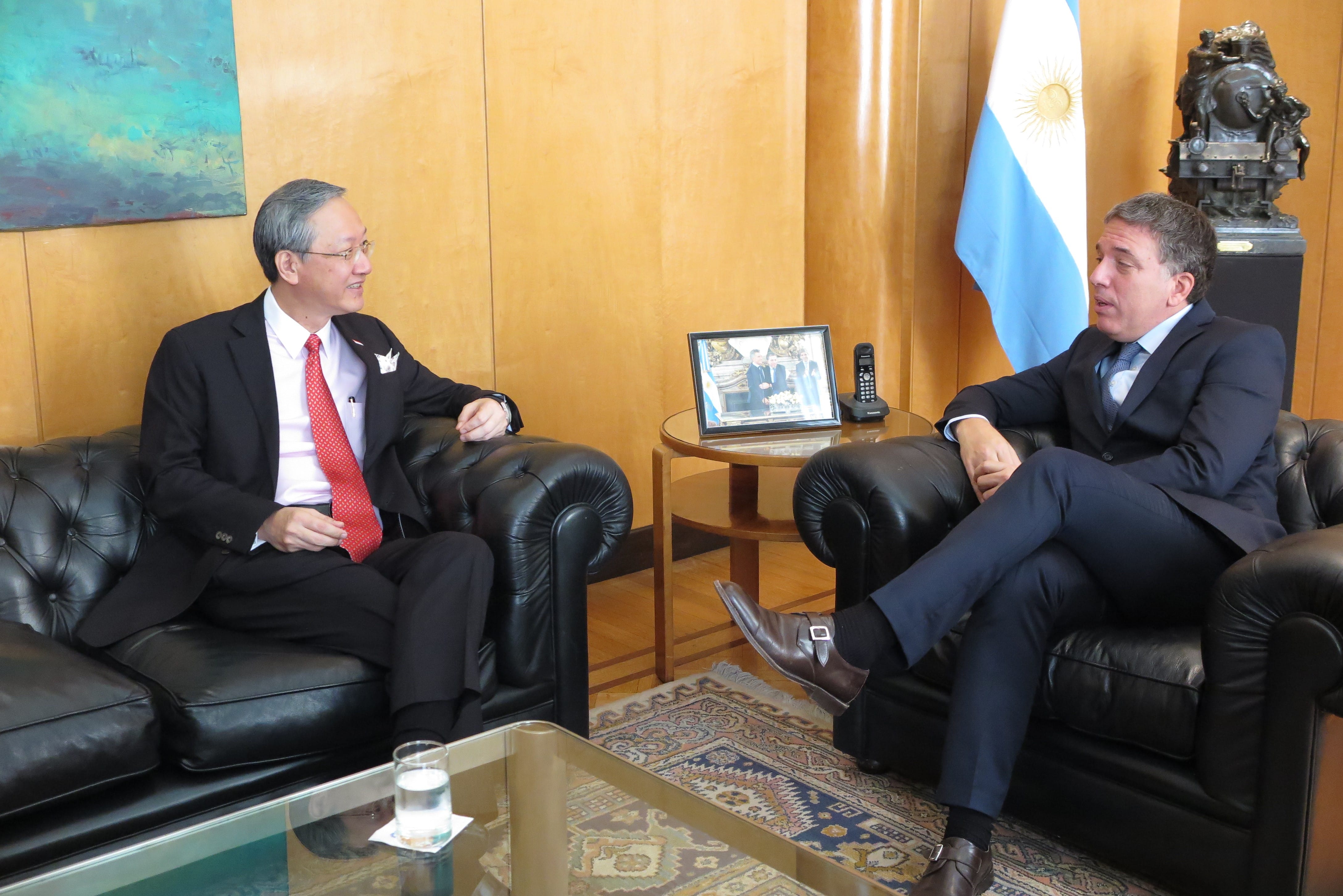 Two men in suits seated in leather chairs with Argentina flag in background.